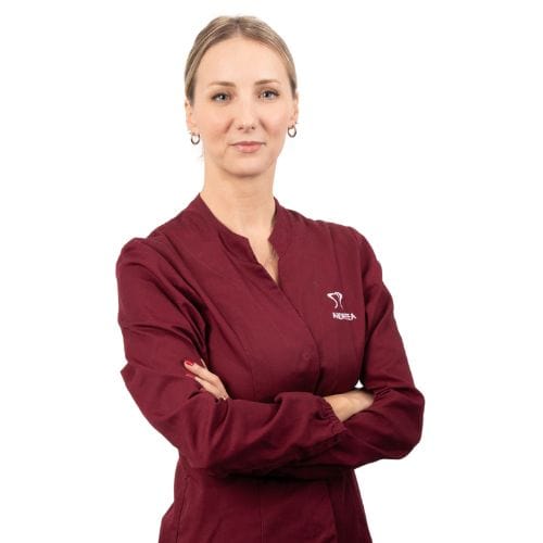 Donna con uno scrub marrone di Dental@Med Group con le braccia incrociate, in piedi su uno sfondo bianco, che guarda la telecamera.
