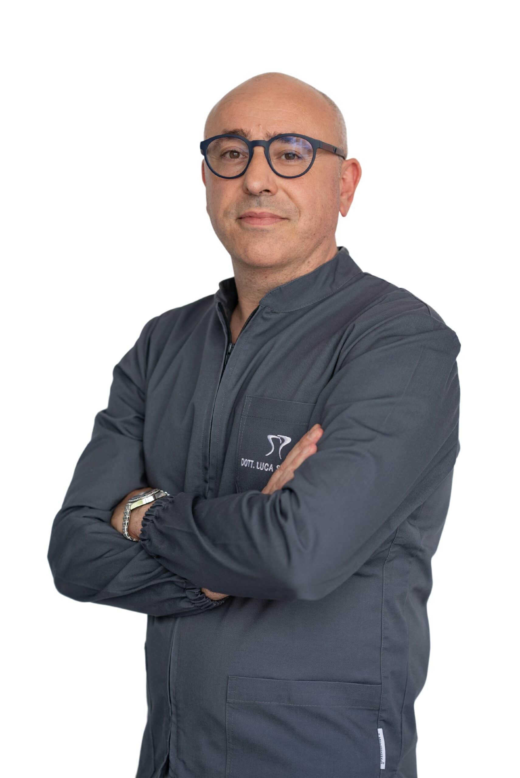 A middle-aged bald man wearing glasses and a gray jacket stands with arms crossed, looking at the camera against a white background, representing Dental@Med Group.