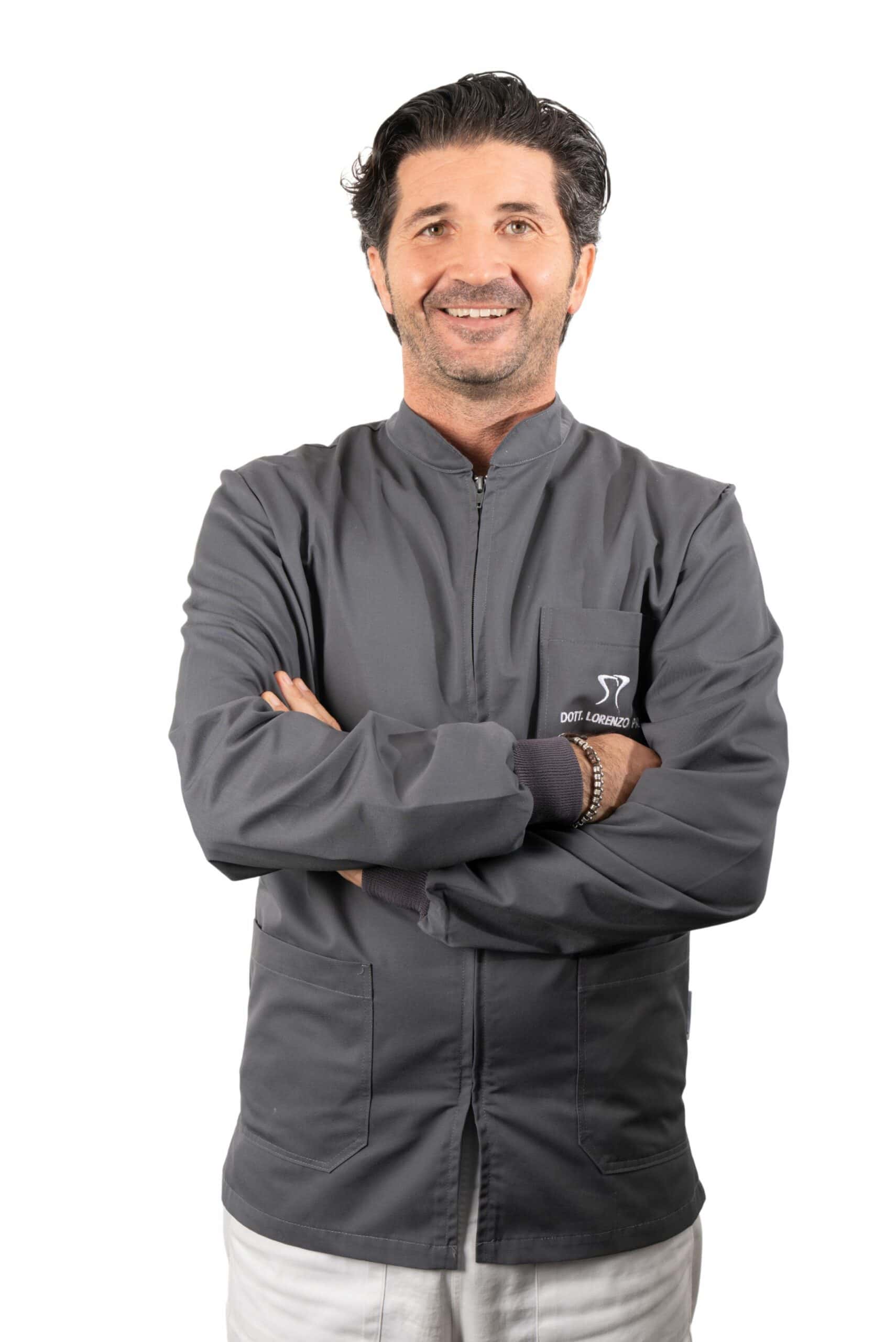 A man with dark hair, wearing a gray jacket and white pants, stands smiling with his arms crossed against a white background. He is a proud member of the Dentalmed Group.