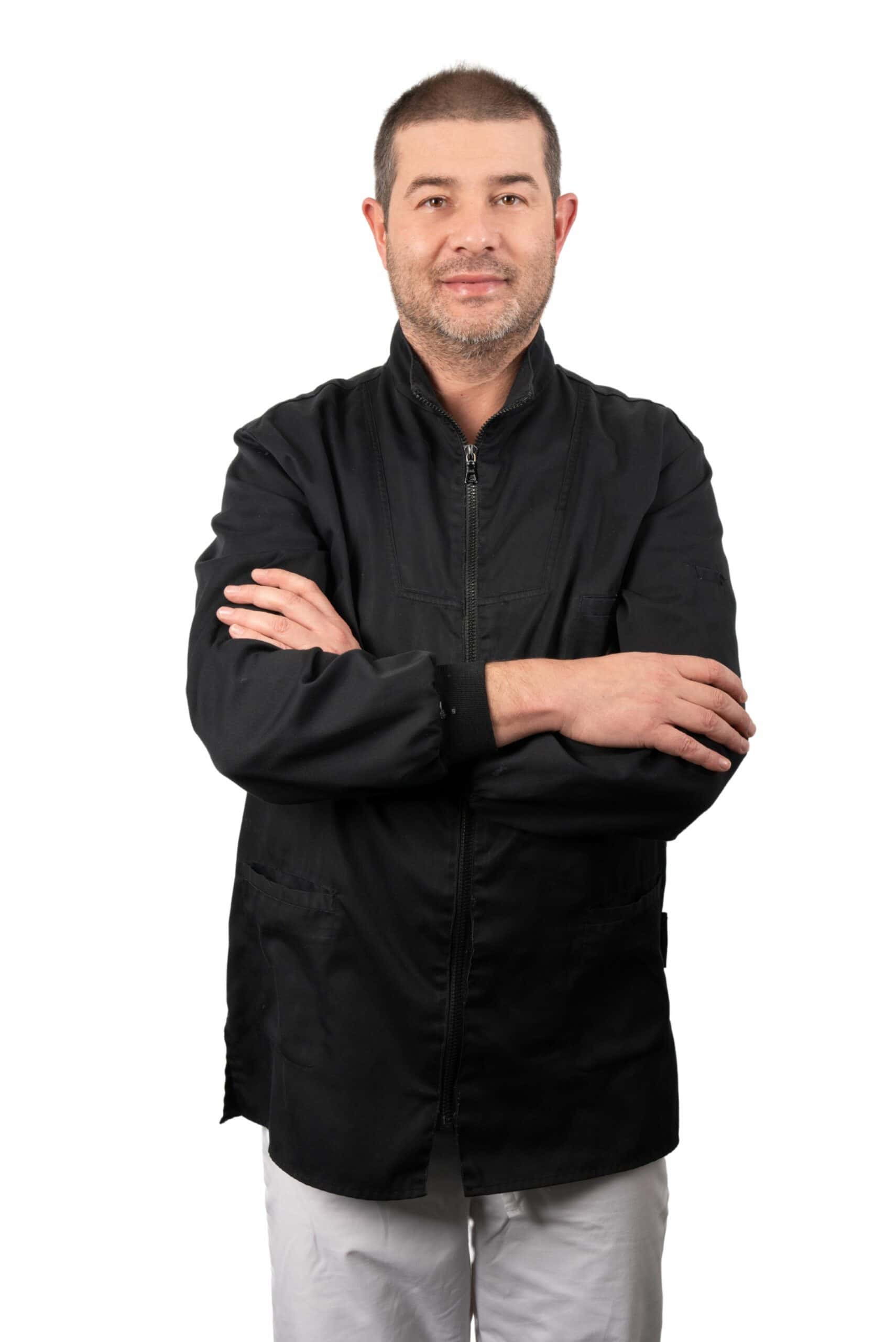 Uomo in giacca nera e pantaloni grigio chiaro in piedi con le braccia incrociate, sorridente alla telecamera, isolata su uno sfondo bianco.