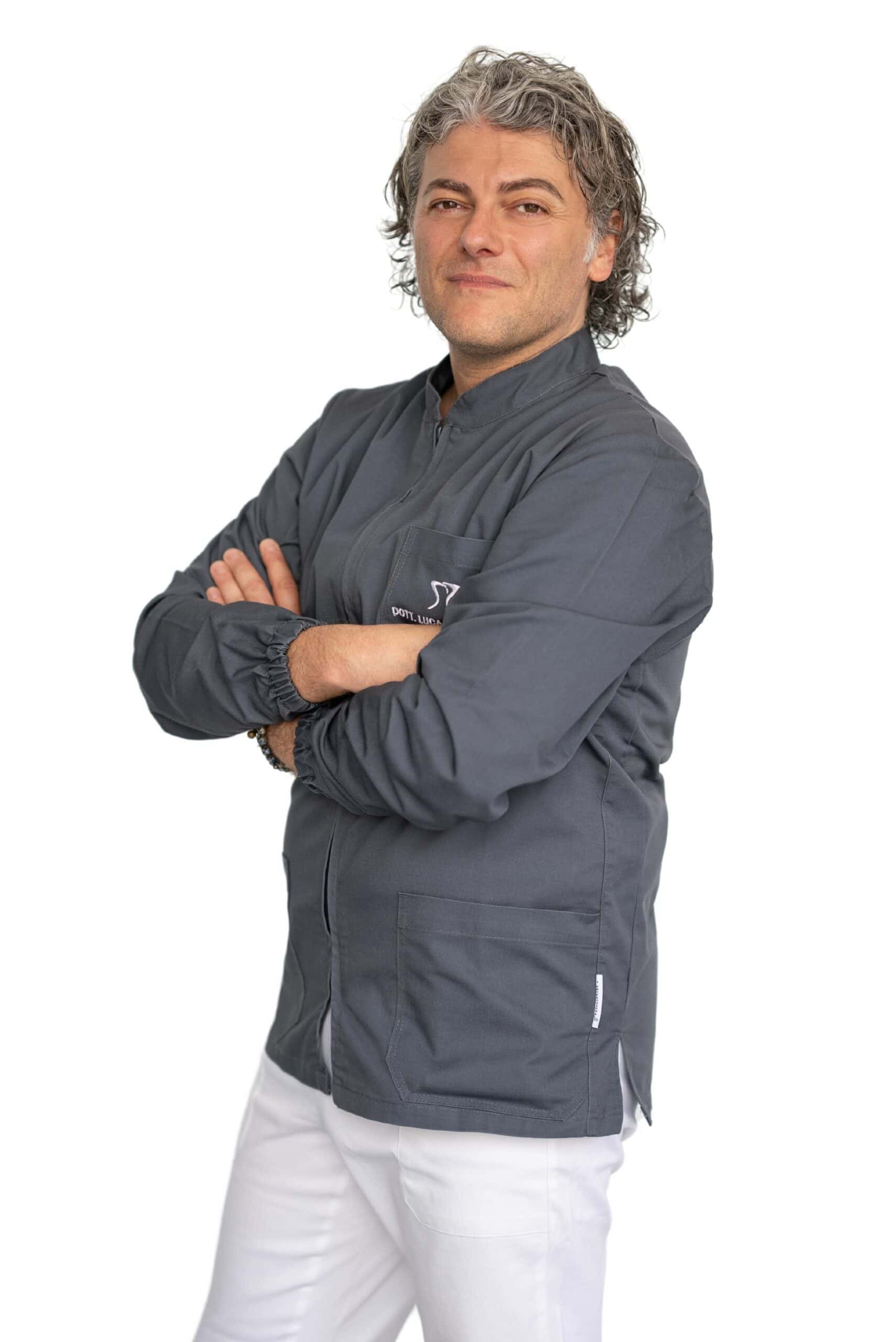 Uno chef fiducioso con i capelli grigi in piedi, le braccia incrociate nella sua giacca da chef grigia e pantaloni bianchi, sorride alla telecamera su uno sfondo bianco.