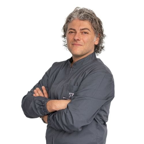 A confident middle-aged man with curly gray hair, wearing a dark jacket from the Dental@Med Group, standing with his arms crossed against a white background.