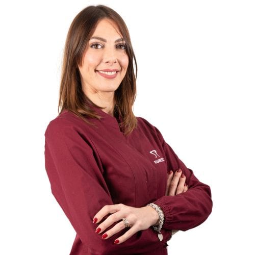 Una donna in divisa medica bordeaux sorride alla telecamera, in piedi con le braccia incrociate presso Dentista Foligno.